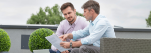 two men discussing a work document together on an outdoor terrace with green spaces and rattan furniture